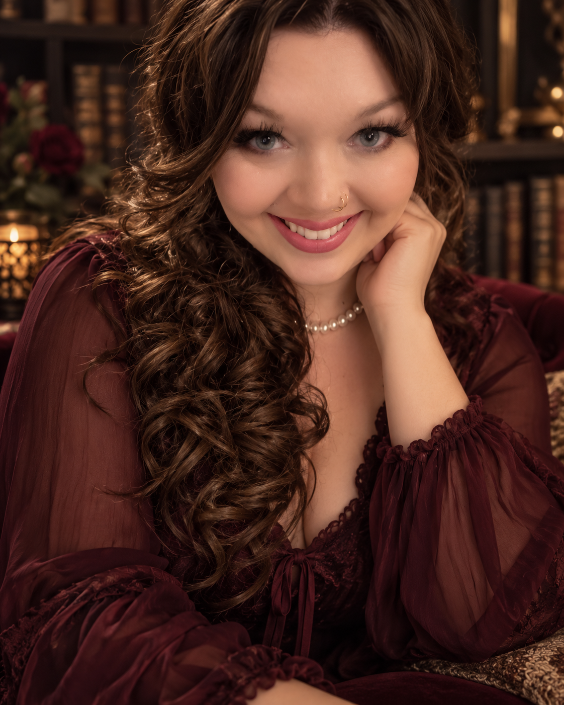 Portrait of author Sloane Devereaux smiling, with long dark curls, a pearl necklace, and a wine-colored sheer blouse, seated before shelves of antique books